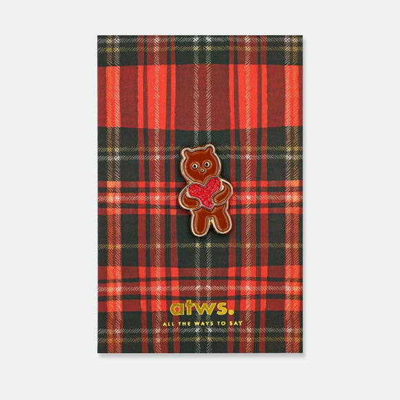 Enamel pin of a brown bear standing upright, holding a red glitter heart with both paws, gold outline, on an illustrated tartan backing card