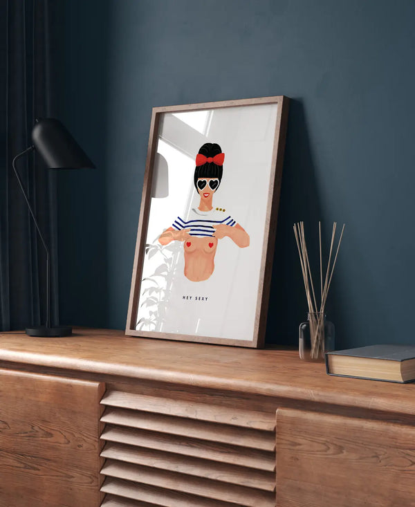 Framed illustration of a woman in a French mariniere top and heart sunglasses on a wooden sideboard against a dark teal wall.