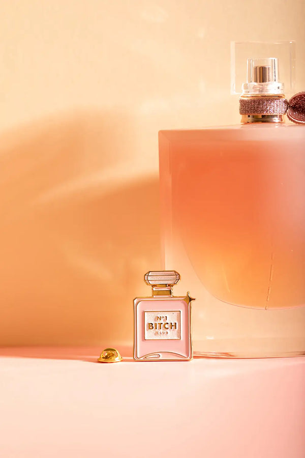 N°1 Bitch - Pins: elegant product shot of the perfume-shaped pin against a warm peach-toned background.