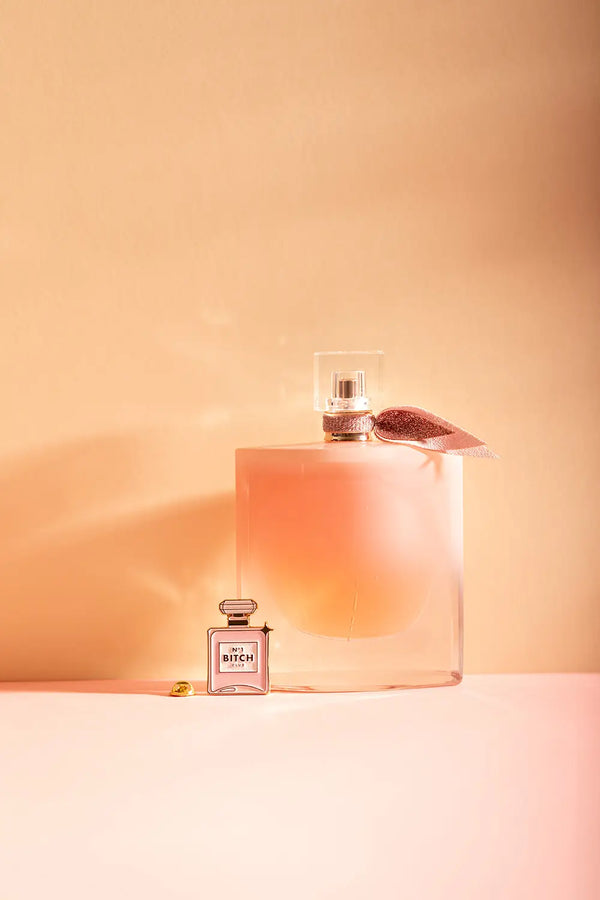 N°1 Bitch - Pins: a pink perfume bottle enamel pin displayed next to a large glass fragrance bottle.