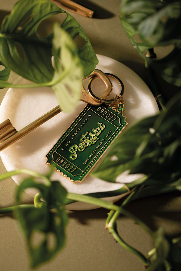 Green "Plant addict ticket club" enamel keychain shaped like a ticket, reading "No space left, one more won't hurt", on a marble dish among monstera leaves