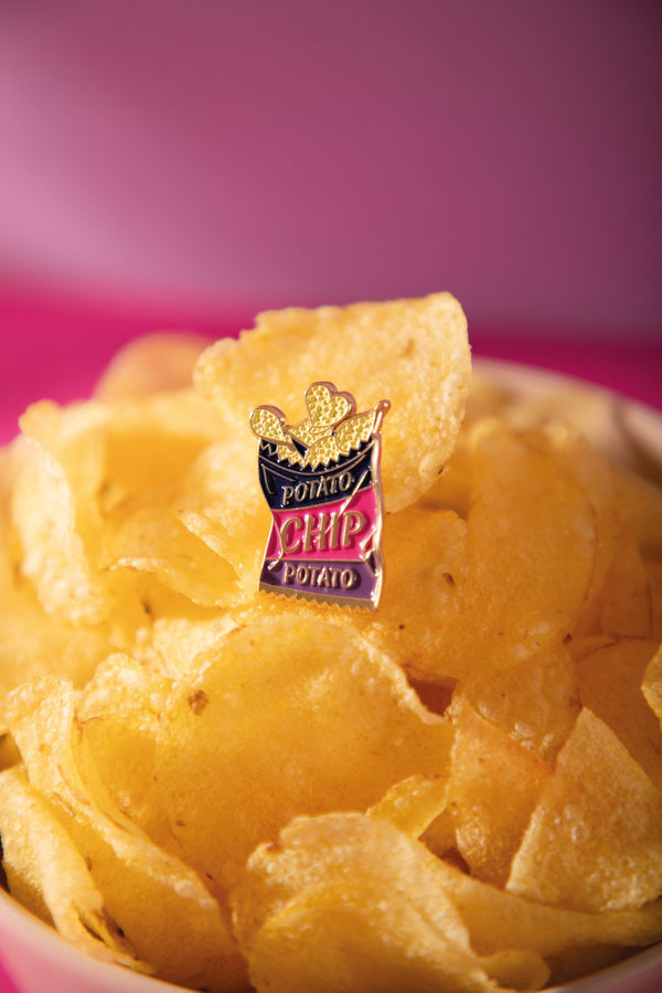 Potato chip bag enamel pin in navy, pink and purple with gold chips, resting on a bowl of crisps