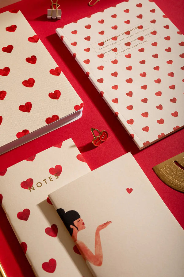 NEW HEARTS - Collection: a stylish flat lay featuring heart-patterned notebooks, a cherry pin, and a woman blowing a heart kiss illustration.