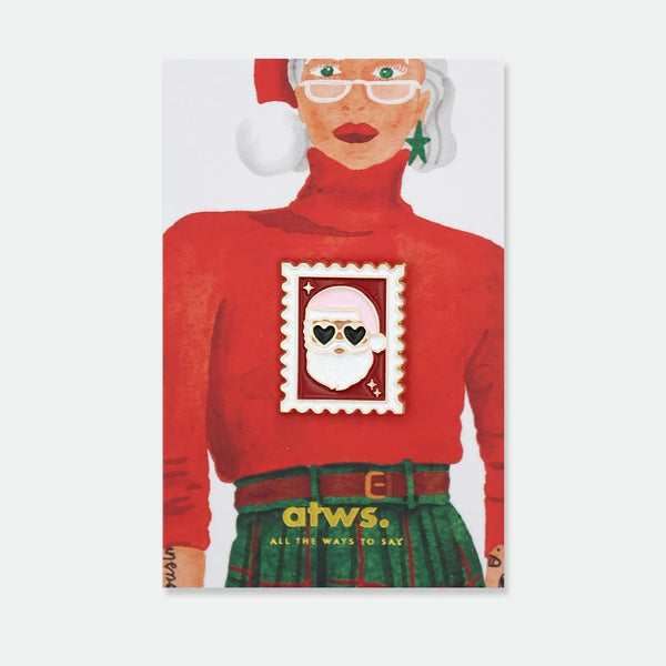 Enamel pin shaped like a postage stamp featuring Santa Claus wearing heart-shaped sunglasses and a glitter beard, gold scalloped border