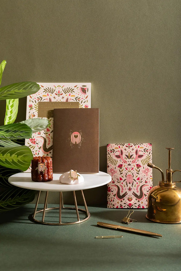 Pink Scarab - Collection: a premium stationery set featuring beetle-illustrated notebooks and enamel pins on a marble stand.