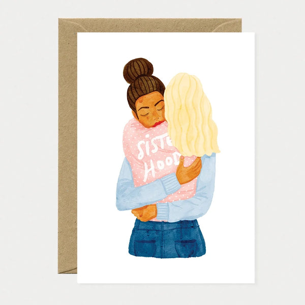 Illustrated greeting card of two women embracing, one wearing a pink sweater with "Sisterhood" written on it and her eyes closed