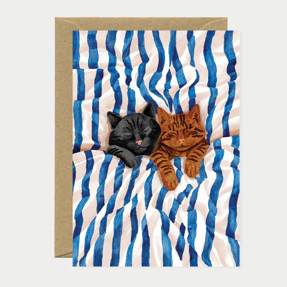 Illustrated greeting card of a black cat and a tabby cat sleeping together on blue and white striped sheets