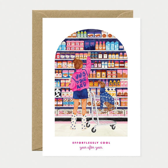 Illustrated birthday greeting card of a woman in a pink jacket reaching for a shelf in a candy and grocery aisle, with a dachshund sitting in the shopping cart below, text reading "Effortlessly cool, year after year"