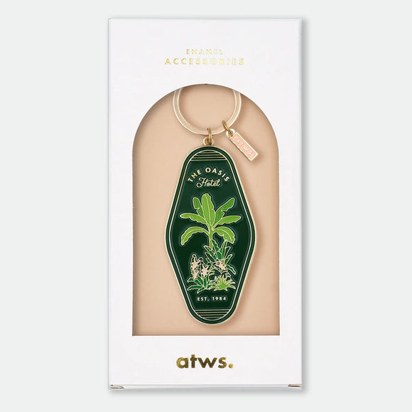 Enamel and metal keychain shaped like a vintage motel key tag, featuring a palm tree and desert flowers with text reading "The Oasis Hotel, Est. 1984," gold-tone ring and pink "ATWS" charm