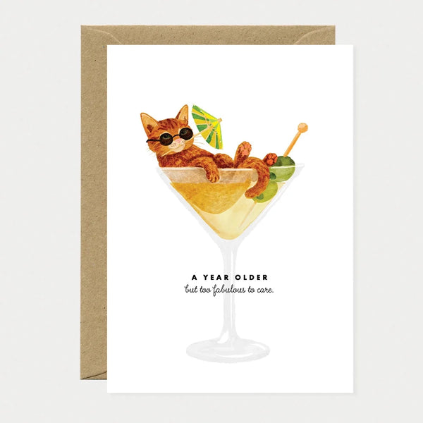 Illustrated birthday greeting card of a ginger cat wearing sunglasses, lounging inside a martini glass with a cocktail umbrella and olives, text reading "A year older, but too fabulous to care"