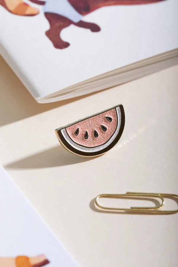 Watermelon - Pins: a sparkling pink glitter watermelon slice enamel pin with gold plating on a cream surface.