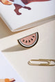 Watermelon - Pins: a sparkling pink glitter watermelon slice enamel pin with gold plating on a cream surface.
