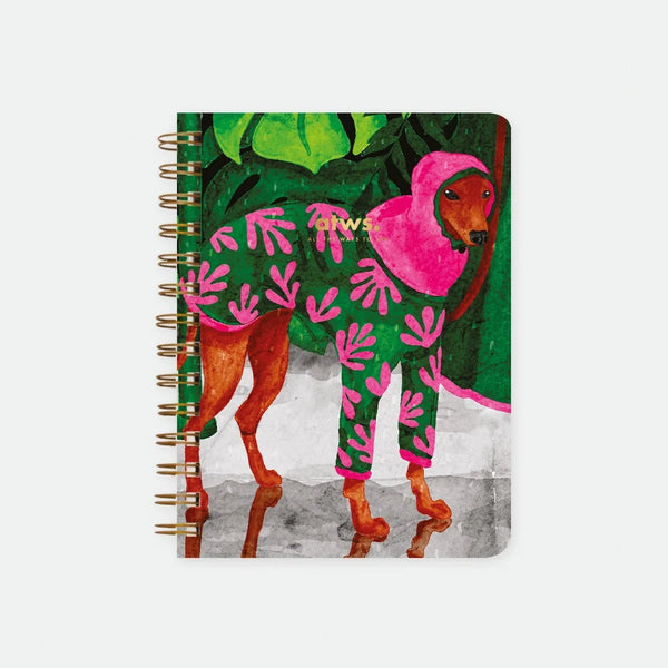 Wet Season A5 spiral notebook by All The Ways To Say, close-up illustration of a dachshund wearing a pink and green leaf-print hooded raincoat standing near a puddle with tropical foliage in the background, spiral binding visible on the left edge